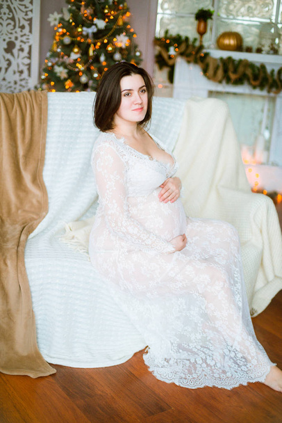 Young pregnant girl with dark hair in a lace white dress on the sofa in a Christmas setting - Photo, Image