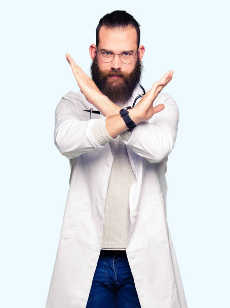Молодий блондин-лікар з бородою в медичному пальто Вираз відторгнення схрещування рук робить негативний знак, розлючене обличчя
 - Фото, зображення