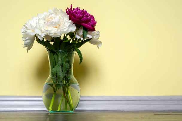 Um vaso de vidro transparente com peônias brancas e rosa fica em um piso cinza contra uma parede amarela. Jarro claro com belo buquê como um presente em um feriado feliz
 - Foto, Imagem