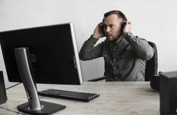 Homme d'affaires ou étudiant confus ou en colère dans une chemise assise contre le moniteur de l'ordinateur dans un casque et parlant avec quelqu'un via Internet. Travailler sur un PC à une table dans le bureau
. - Photo, image