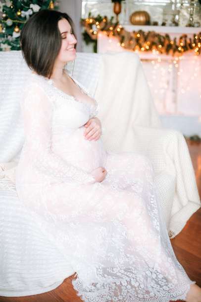 Young pregnant girl with dark hair in a lace white dress on the sofa in a Christmas setting - Foto, imagen