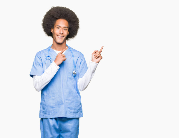 Jovem médico afro-americano homem com cabelo afro sorrindo e olhando para a câmera apontando com duas mãos e dedos para o lado
. - Foto, Imagem