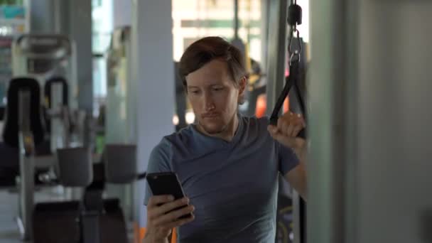 A young man at the gym doing exercises holding a phone in front of his eyes in his hands. All the attention is in the phone. The concept of dependence on social networks. Mobile addiction concept - Materiał filmowy, wideo