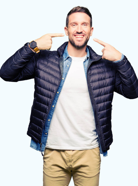 Homem bonito usando casaco de inverno sorrindo confiante mostrando e apontando com os dedos dentes e boca. Conceito de saúde
. - Foto, Imagem