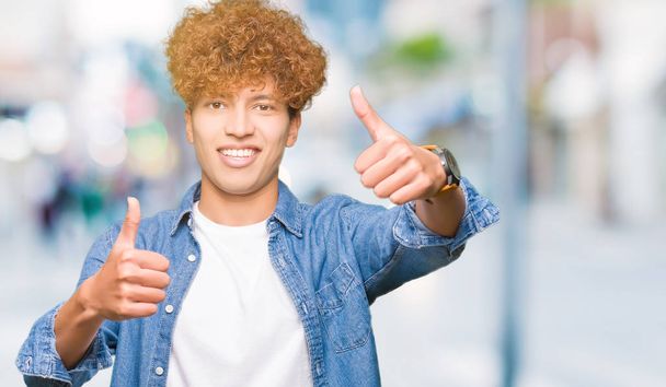 Young handsome man with afro hair wearing denim jacket approving doing positive gesture with hand, thumbs up smiling and happy for success. Looking at the camera, winner gesture. - Photo, Image