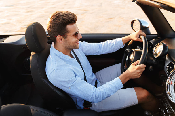 Photo of happy caucasian man driving convertible car by seaside  - 写真・画像