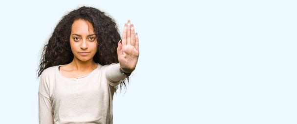 Mooi meisje met krullend haar het dragen van casual trui stop doen zingen met de palm van de hand. Waarschuwing expressie met negatieve en ernstige gebaar op het gezicht. - Foto, afbeelding