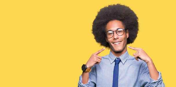 Joven hombre de negocios afroamericano con el pelo afro usando gafas sonriendo confiado mostrando y señalando con los dedos los dientes y la boca. Concepto de salud
. - Foto, imagen