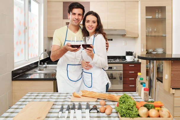 Молода кавказька пара в фартухів п'ють червоне вино при приготуванні страв французької кухні в домашніх умовах - Фото, зображення