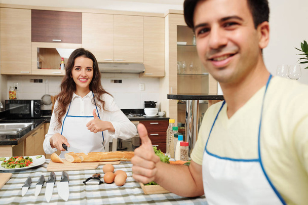 Positiivinen nuori valkoihoinen aviomies ja vaimo ruoanlaitto illallinen yhdessä keittiössä ja näyttää peukalot ylöspäin
 - Valokuva, kuva