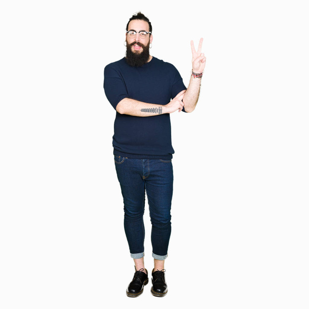 Jonge hipster man met lange haren en baard dragen van een bril glimlachend met blij gezicht winking op de camera doen overwinning teken. Nummer twee. - Foto, afbeelding