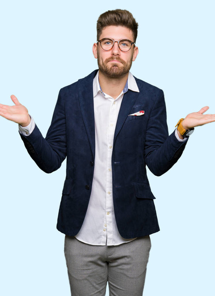 Young handsome business man wearing glasses clueless and confused expression with arms and hands raised. Doubt concept. - Photo, Image