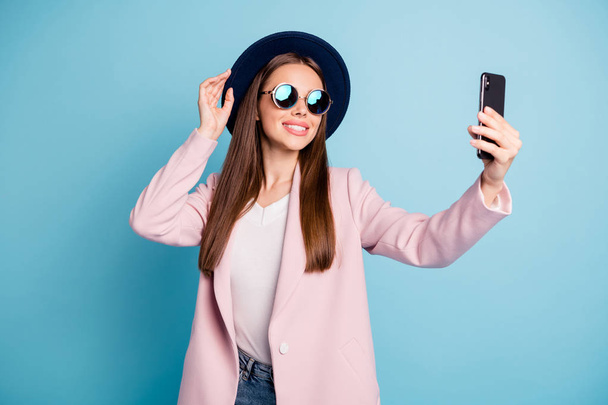 Portrit of sweet lovely girl using her cell phone making selfie posing touching her fashionable hat wearing pink topcoat isolated over blue background - Photo, image