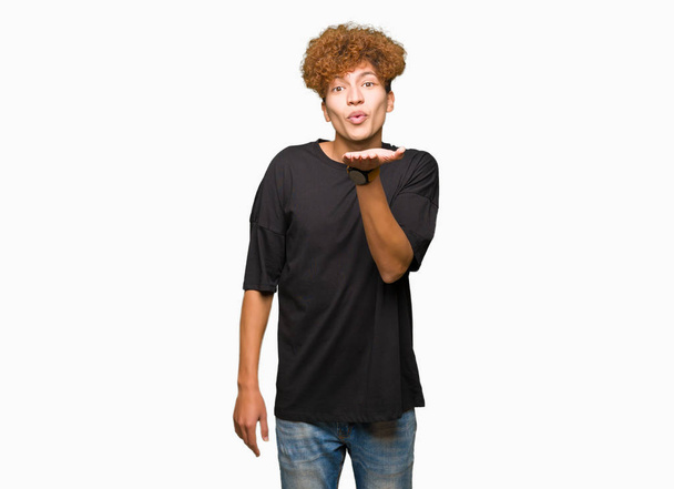 Young handsome man with afro hair wearing black t-shirt looking at the camera blowing a kiss with hand on air being lovely and sexy. Love expression. - Photo, Image