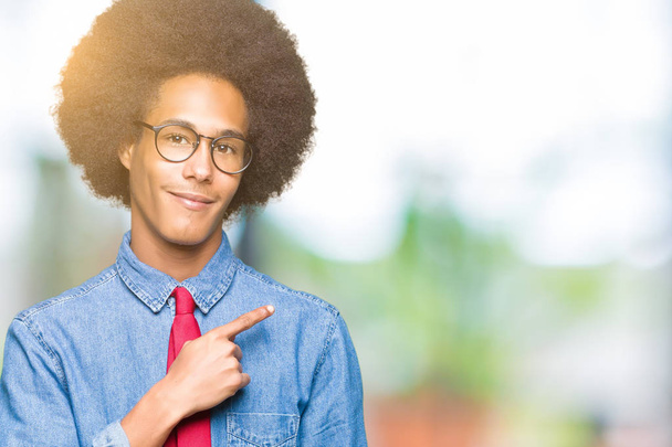 Молодой африканский бизнесмен с афроволосами в очках и красном галстуке веселый с улыбкой на лице, указывающей рукой и пальцем в сторону со счастливым и естественным выражением лица
 - Фото, изображение