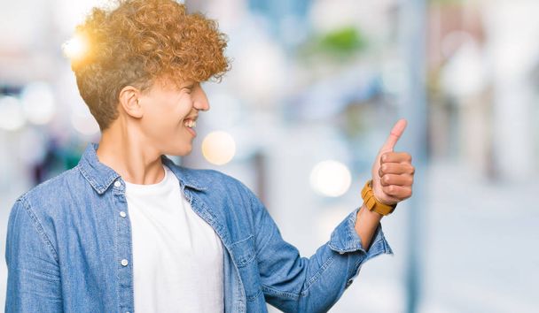 Young handsome man with afro hair wearing denim jacket Looking proud, smiling doing thumbs up gesture to the side - Photo, Image