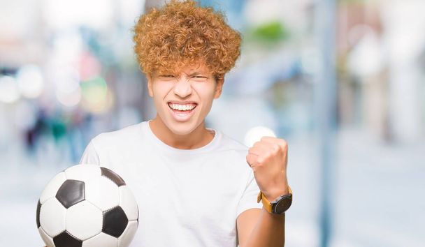 Νέος όμορφος άνθρωπος κρατώντας ποδόσφαιρο μπάλα ποδόσφαιρο φωνάζοντας υπερήφανος και γιορτάζοντας τη νίκη και την επιτυχία πολύ ενθουσιασμένοι, επευφημίες συγκίνηση - Φωτογραφία, εικόνα