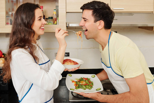 Happy young woman in love feeding her boyfriend with spaghetti when they are standing in kitchen after cooking dinner - Photo, Image