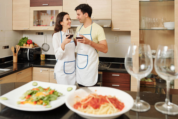 Θετικό νεαρό ζευγάρι σε ποδιές που στέκονται στο πάγκο της κουζίνας με ποτήρια κρασιού στα χέρια και κοιτάζοντας ο ένας τον άλλο με αγάπη - Φωτογραφία, εικόνα