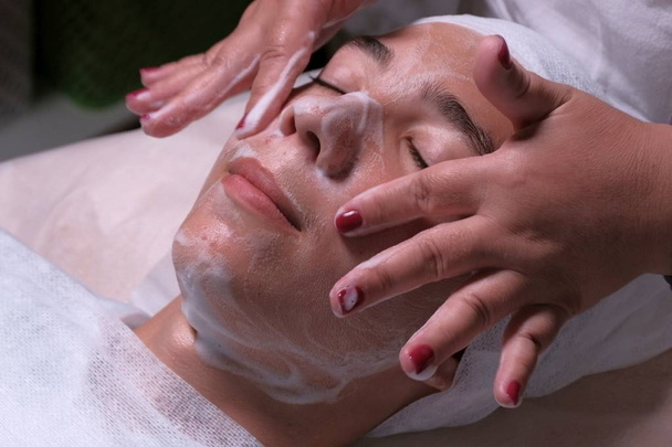 Teenager in beauty clinic. Close-up of the hands of a beautician who lathers the face of a boy. Male client gets facial cosmetology procedure in beauty salon. Medical face skin acne cleaning. - Photo, Image