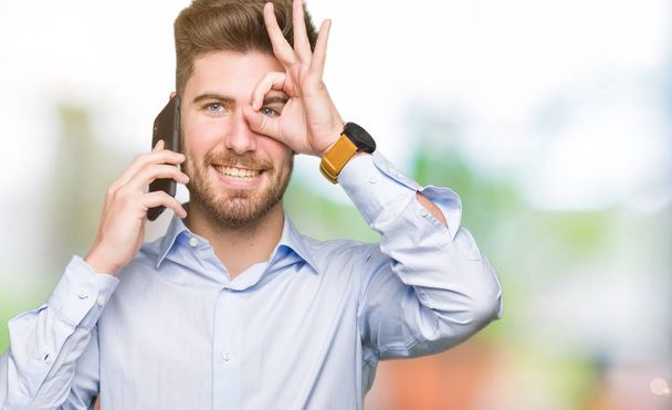 Молодой красавчик разговаривает по смартфону со счастливым лицом, улыбается, делает знак "ОК" с рукой на глазу, глядя сквозь пальцы
 - Фото, изображение