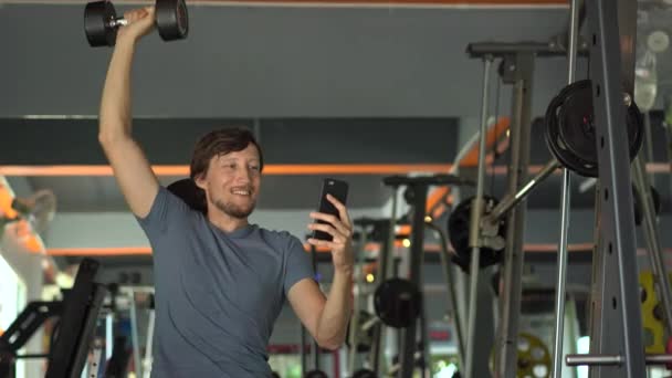 A young man at the gym doing exercises holding a phone in front of his eyes in his hands. All the attention is in the phone. The concept of dependence on social networks. Mobile addiction concept - Záběry, video