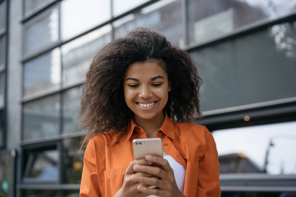 Portret Wesoła African American kobieta za pomocą aplikacji mobilnej na zakupy online. Udane blogger blogu w sieciach społecznościowych. Uśmiechnięta dziewczyna trzyma smartphone, rozmowy, rezerwacje biletów - Zdjęcie, obraz