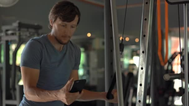 A young man at the gym doing exercises holding a phone in front of his eyes in his hands. All the attention is in the phone. The concept of dependence on social networks. Mobile addiction concept - Imágenes, Vídeo