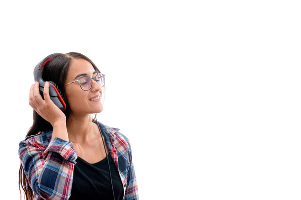 Kaukaska młoda dziewczyna z koszulą i okularami, słuchanie muzyki z dużymi słuchawkami i telefonem komórkowym lub innego rodzaju urządzenia elektronicznego na białym tle izolowane - Zdjęcie, obraz