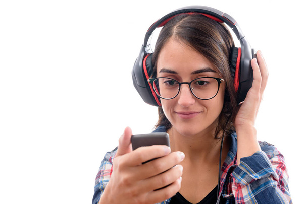 Καυκάσιο νεαρό κορίτσι με καρό πουκάμισο και γυαλιά, ακούγοντας μουσική με μεγάλα ακουστικά και ένα κινητό τηλέφωνο ή άλλο τύπο ηλεκτρονικής συσκευής σε λευκό φόντο απομονωμένη - Φωτογραφία, εικόνα