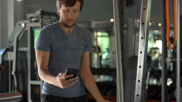 A young man at the gym doing exercises holding a phone in front of his eyes in his hands. All the attention is in the phone. The concept of dependence on social networks. Mobile addiction concept - Felvétel, videó