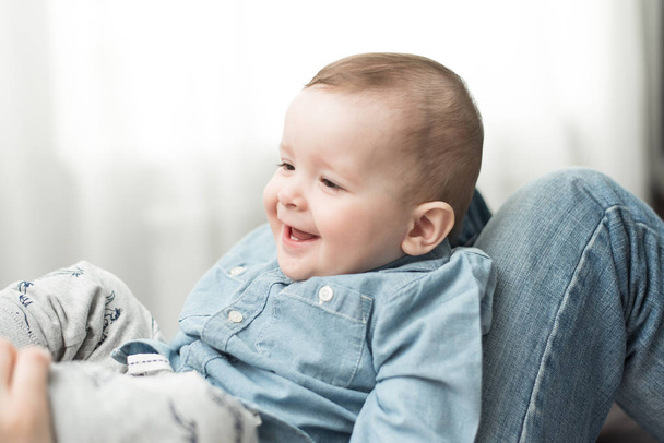 ένα μωρό ενός έτους κάθεται στην αγκαλιά της μάνας του και γελάει. Ένα μικρό ξανθό αγόρι χαμογελά σε ένα κοστούμι τζιν σε ελαφρύ φόντο. - Φωτογραφία, εικόνα