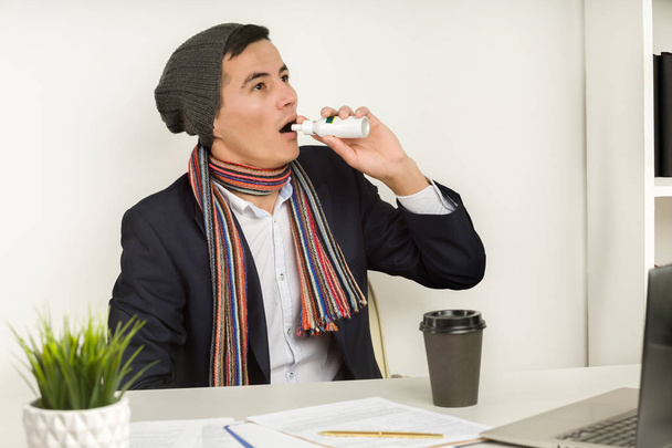Aziatische man in een hoed, sjaal en jas maakt gebruik van een spray in Office omdat gebroken verwarming of koeling van de airconditioner overmatig. keelpijn of ontsteking van de amandelen - Foto, afbeelding