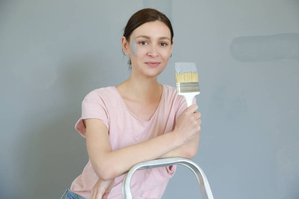 Портрет молодой женщины, держащей кисть, женщина рисует серой краской на белой стене
 - Фото, изображение