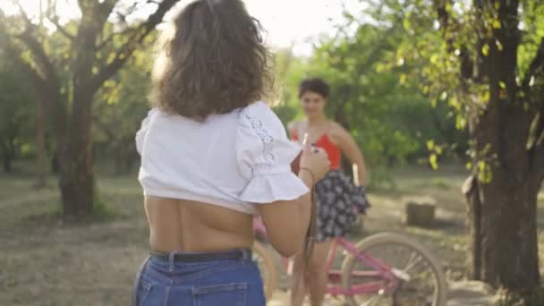 Cute young caucasian woman with short hair posing with her bicycle while her friend taking a photo in the garden or park. Rural life. Retro style - Πλάνα, βίντεο