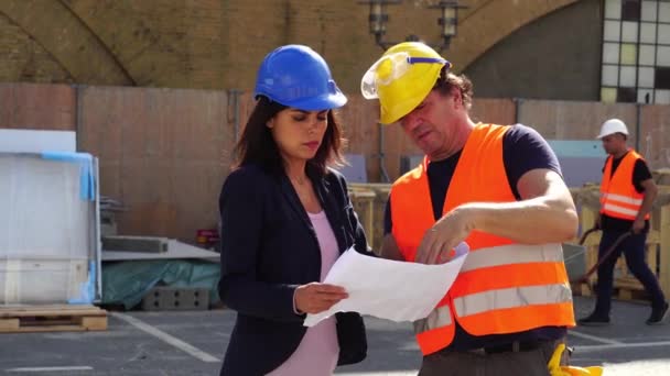 Ingenieros civiles masculinos y femeninos que llevan chalecos de protección, googles y gorras duras revisando proyectos y planos de oficinas en el sitio de construcción
 - Metraje, vídeo