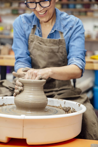 Goed uitziende Senior vrouwelijke Potter werken op aardewerk wiel terwijl zittend in haar workshop - Foto, afbeelding