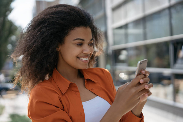 Portrait of successful blogger holding smartphone, streaming video online, writing blog post outdoors. Cheerful African American woman using mobile phone, chatting, writing text message, communication - Foto, Imagen