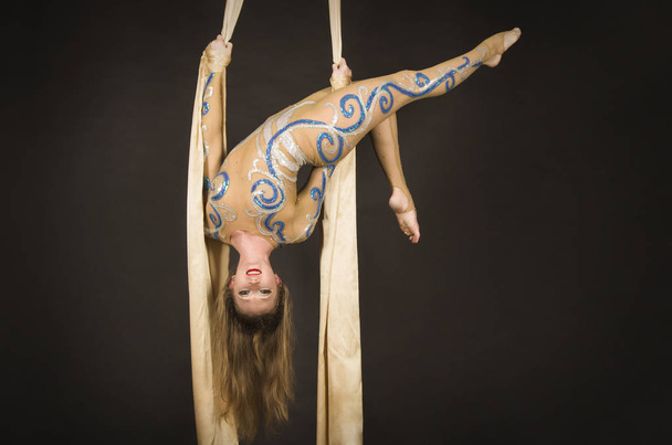 A slender girl in a beige suit performs gymnastic and circus exercises on Golden silk. - Foto, afbeelding