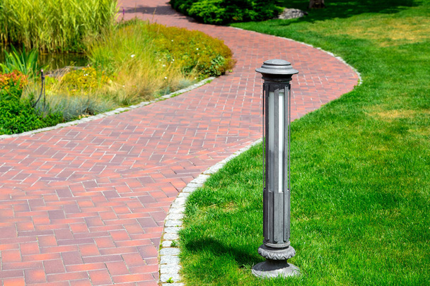 ground street lamp mounted on a green lawn in a park with plants and a winding curved footpath to a pond with a flower bed and reeds on sunny day. - Photo, Image