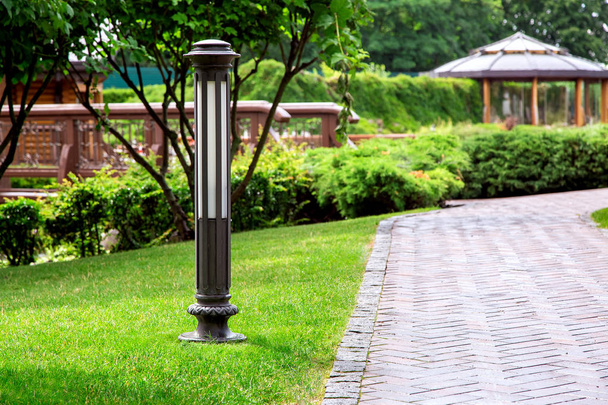 Iron ground lantern mounted in the green lawn of the backyard with footpath of stone tiles among green spaces, on a summer day. - 写真・画像