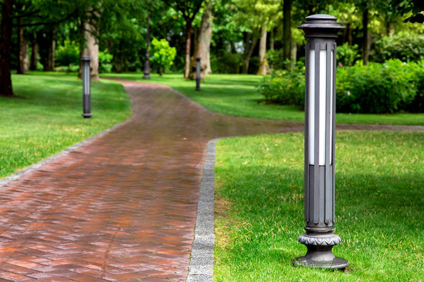 Iron ground garden lantern installed in the green lawn of the park with footpath of red stone tiles among green spaces, on a summer day after rain nobody. - Фото, изображение