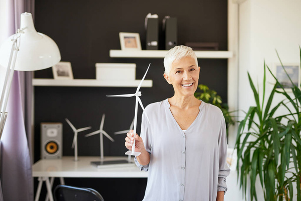 Beautiful smiling Caucasian senior woman standing in office and holding windmill model. Start up business concept. - Photo, Image
