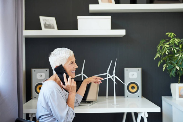 Linda mulher idosa caucasiana falando no telefone e tocando modelo de moinho de vento enquanto sentado no escritório. Iniciar conceito de negócio
. - Foto, Imagem