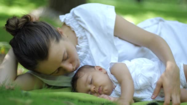 Mamá besa a su bebé durmiendo en una hierba verde al aire libre. Feliz y sonriente madre e hijo en el Green Summer Park. Hermosa familia en el parque de primavera disfrutando de la naturaleza
. - Imágenes, Vídeo