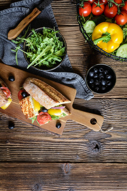 Wholemeal vegetarian baguette stuffed with grilled white cheese, arugula, tomatoes, black olives, bell pepper and red onion - Photo, Image