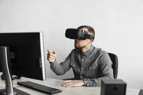 Young bearded man sitting against computer with virtual reality glasses trying to touch something or tap some thing in front of what he seeing. Modern technologies. The concept of future technology. - Foto, Imagen