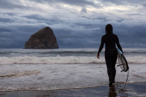Avontuurlijk meisje met een surf plank gaat surfen in de Oceaan tijdens een bewolkte zomer zonsondergang. Genomen in Pacific City, Oregon Coast, Verenigde Staten van Amerika. - Foto, afbeelding