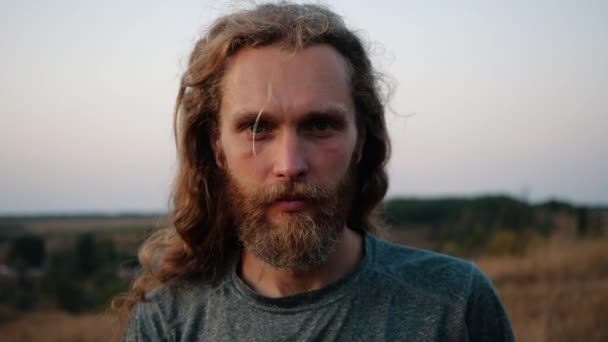 Közelről, portré egy csinos kaukázusi szakállas fiatal férfi, hosszú áramló hajat keres kedvesen a kamera a háttérben egy fényes naplemente felett természet. Élet öröme, aktív életmód. - Felvétel, videó