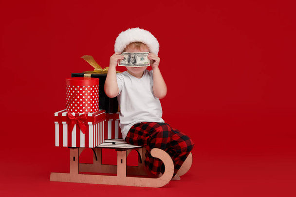 Un petit garçon dans un chapeau de Père Noël est assis sur un traîneau en bois avec des boîtes-cadeaux et tient un billet de banque. Le concept de Noël et Nouvel An Cashback. Réductions et ventes
 - Photo, image